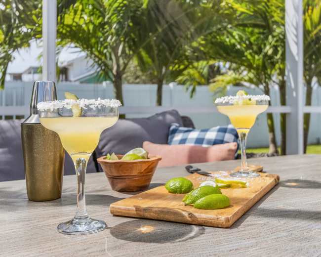 Two margarita cocktails with salt-rimmed glasses sit on a table alongside a wooden cutting board with limes and a cocktail shaker, set against a backdrop of palm trees.