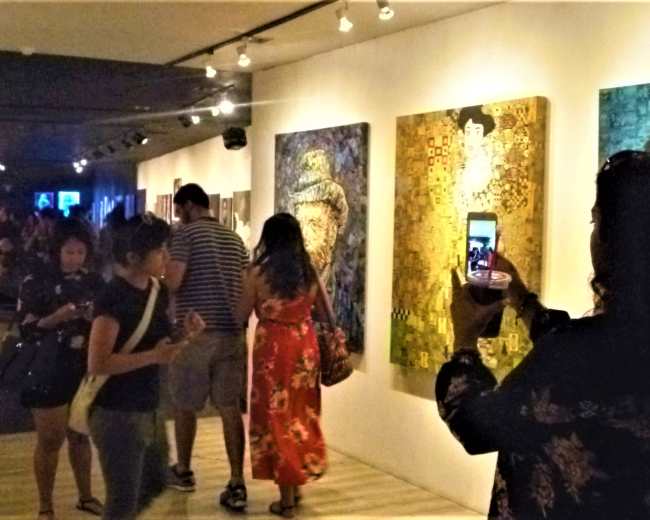 A group of visitors explores an art gallery, with some individuals viewing paintings while others take photos.