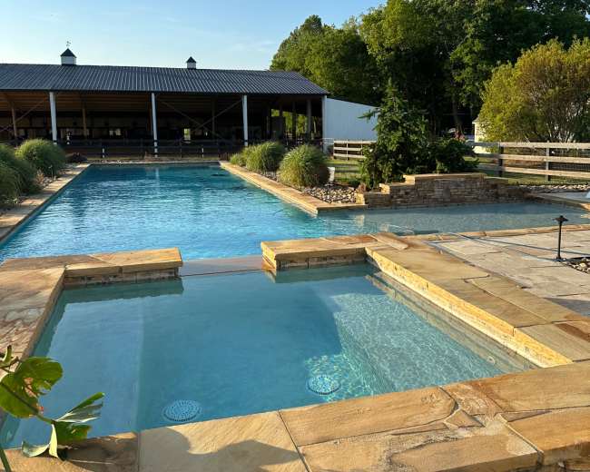A clear outdoor swimming pool with a spa area, surrounded by stone coping and greenery, under a blue sky with a few clouds.