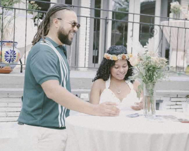 A man with dreadlocks and sunglasses stands beside a woman in a floral crown at a round table in an outdoor setting.
