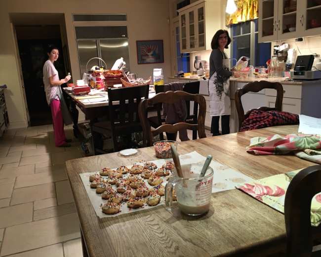 Two women are preparing cookies in a brightly lit kitchen, with a tray of cookies and various baking supplies spread across the countertop and table.