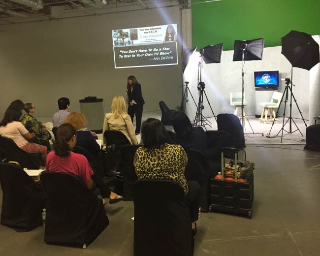 A speaker presents to an audience in a studio setup with lighting equipment and a television screen in the background.