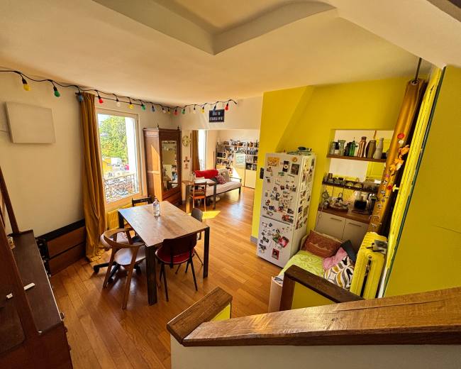 The image shows a bright, open kitchen and living area with wooden flooring, yellow walls, and a dining table surrounded by chairs, along with decorative string lights.