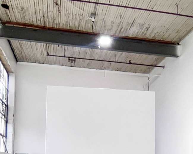 The image shows a large, empty white canvas on wheels in an industrial-style art studio.