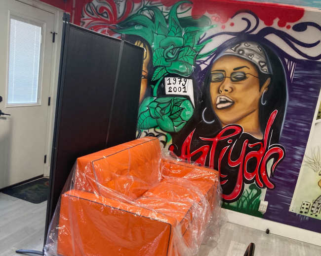 A bright orange sofa wrapped in plastic is positioned next to a partition in a room featuring a colorful mural on the wall.