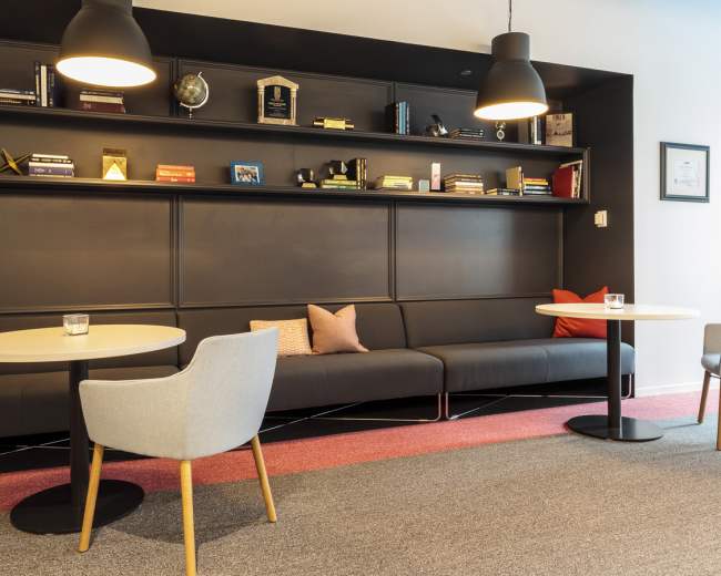 The image shows a modern lounge area with a dark shelving unit displaying various items, two round tables, and comfortable seating with pillows.