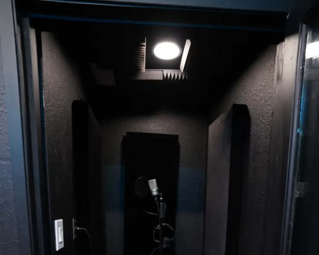 The image shows a small, soundproof recording booth with a microphone on a stand and acoustic panels on the walls.
