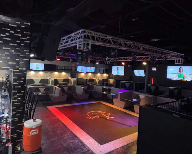 The image shows an empty nightclub with seating areas, multiple screens on the walls, and colorful lighting effects on the floor.