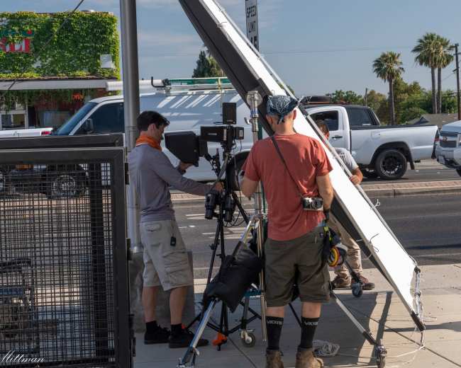 A film crew sets up equipment on a sidewalk near a restaurant and palm trees under clear skies.