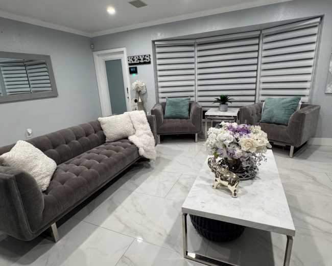 The image shows a modern living room with a gray tufted sofa, two armchairs with cushions, a marble coffee table, and floral decorations.