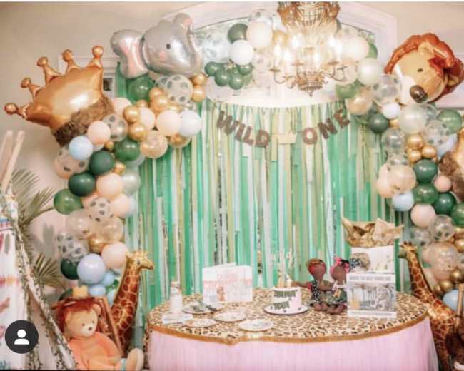 A colorful party setup features balloon decorations, a table with themed items, and greenery for a "Wild One" celebration.