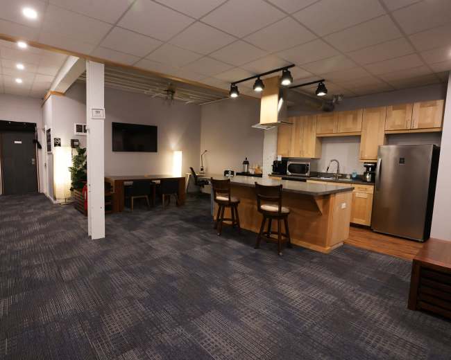 The image shows a modern kitchen and dining area with wood cabinetry, a countertop, and a small table with chairs, adjacent to a hallway.