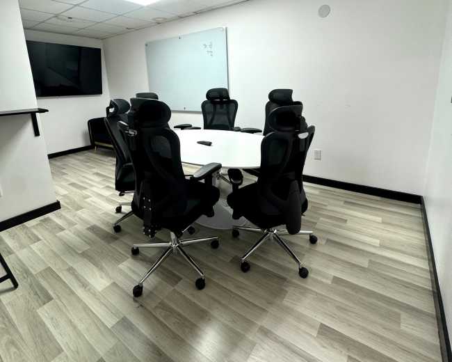 The image shows a conference room with a round table surrounded by six ergonomic chairs, a large TV on one wall, and a whiteboard on another.