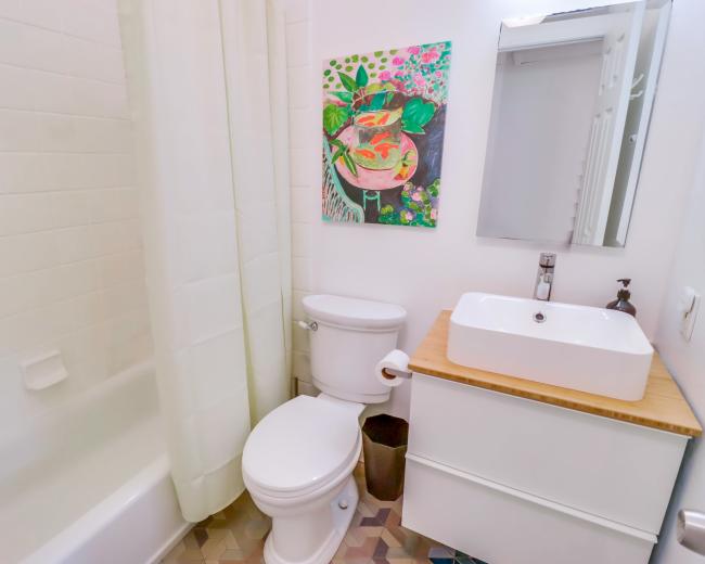 The bathroom features a bathtub with a shower curtain, a toilet, a wall-mounted sink, and a colorful painting above the sink.