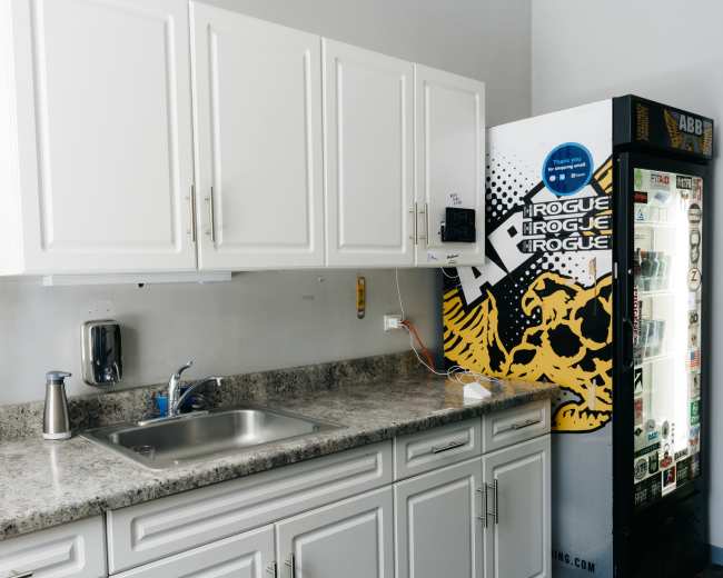 The image shows a kitchen area featuring white cabinets, a stainless steel sink, and a vending machine against a wall with a graphic design.
