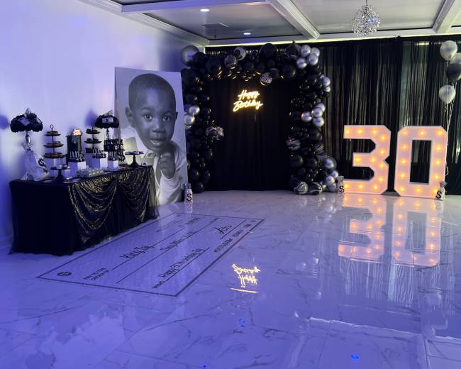 A decorated indoor venue for a 30th birthday celebration, featuring a dessert table, a large black and white portrait, and illuminated "30" balloons.
