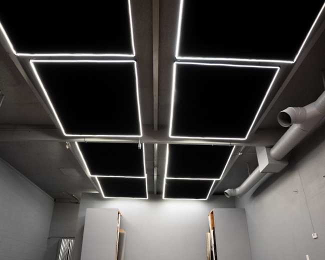 An empty room features a gray wall, a bare concrete floor, and rectangular light fixtures mounted on the ceiling.