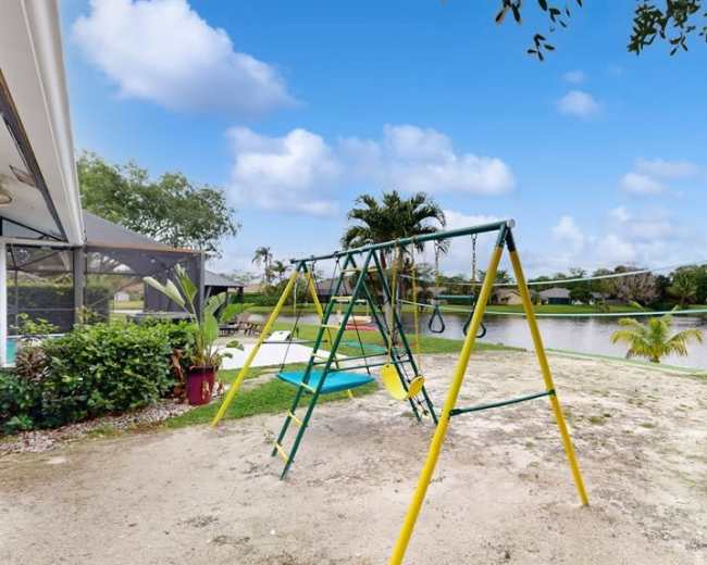 A playground swing set stands on a grassy area next to a calm lake with palm trees in the background.