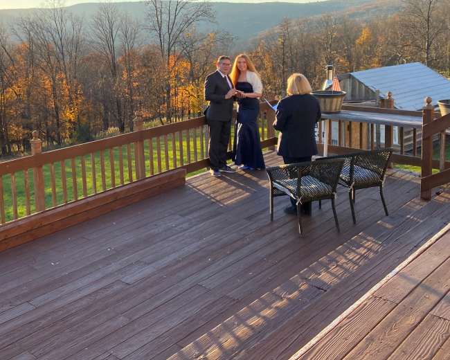 Three people stand on a wooden deck overlooking a landscape of trees with autumn foliage and rolling hills.