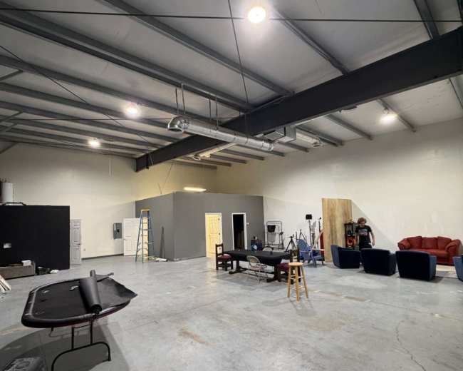 The image shows an industrial-style studio space with concrete floors, metal beams, and various pieces of furniture, including a couch and chairs, arranged in the center.
