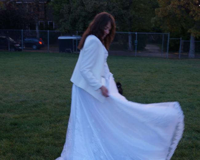 A woman in a white wedding dress twirls on green grass as the sun sets behind her.