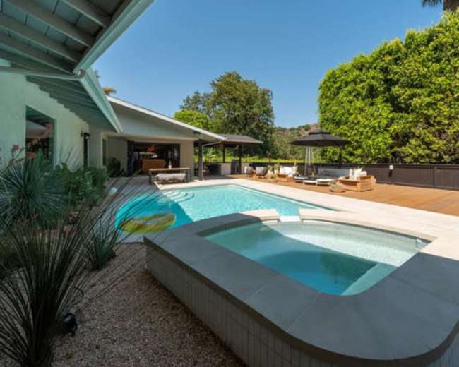 The image shows a modern outdoor space featuring a swimming pool and a hot tub surrounded by lounge chairs and greenery.