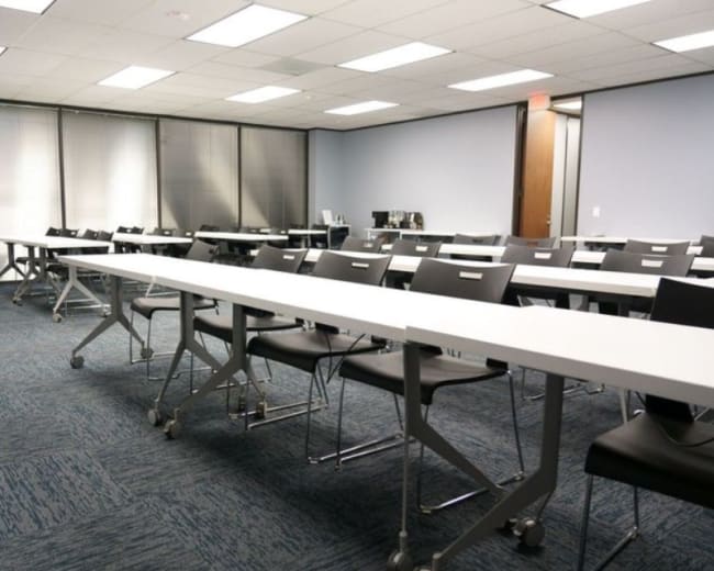 The image shows a modern classroom with rows of white tables and black chairs arranged for a presentation, alongside a podium in the corner.