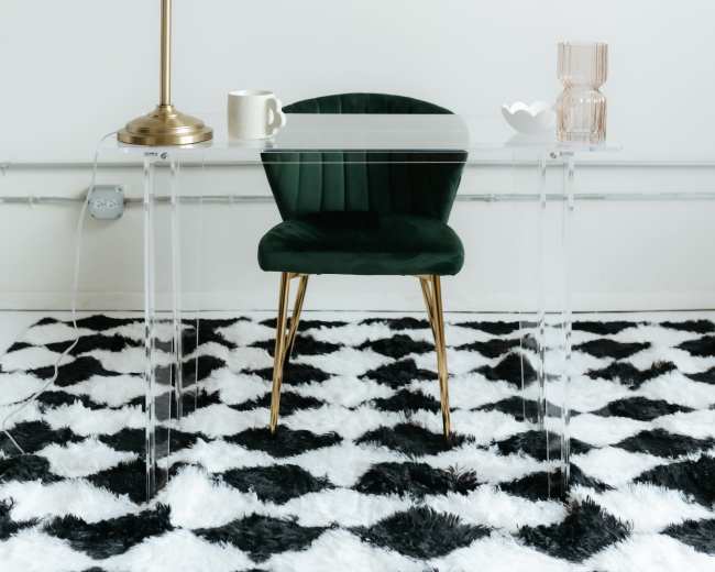 A clear glass desk with a green chair, a gold lamp, a white mug, and a decorative vase sits on a black and white striped rug.