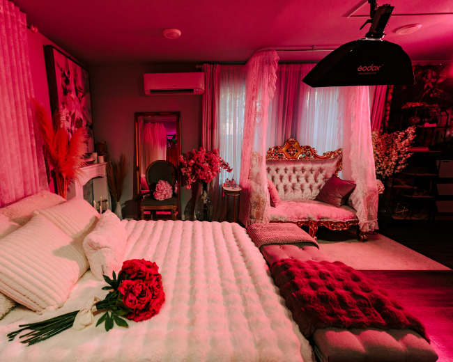 The image shows a styled bedroom featuring a plush bed with a white coverlet, a bouquet of red flowers on the bed, and an ornate seating area with decorative elements.