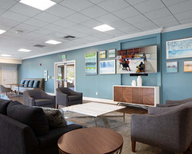 The image shows a modern lounge area featuring gray sofas and armchairs arranged around a coffee table, with a television mounted on the wall displaying a sports scene, and colorful artwork hanging on the blue walls.