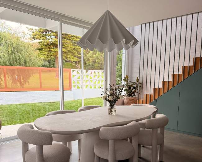 A modern dining area features a round table surrounded by four rounded chairs, with a pendant light hanging above and a view of a green outdoor space through large glass windows.