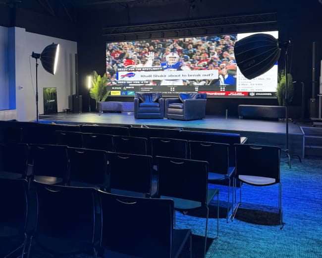 A modern studio setting features a large screen displaying a sports event, with two chairs positioned in front and camera equipment set up in the foreground.
