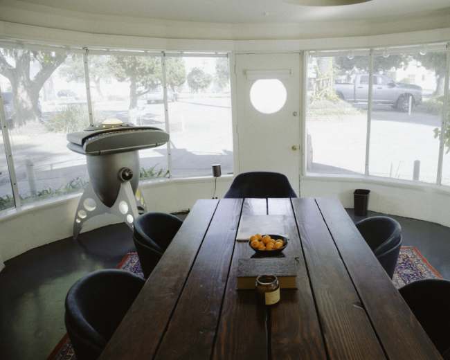 A circular dining area features a large wooden table surrounded by dark chairs, with a round window and a visible outdoor scene.