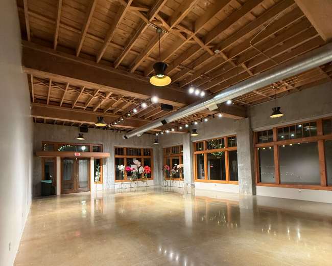 The image depicts a spacious, empty room with polished concrete floors, exposed wooden beams, and large windows.