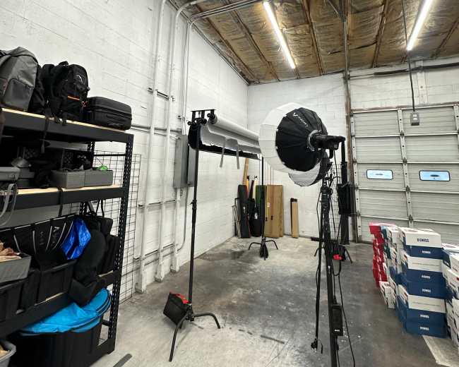 A well-lit photography studio features a seamless backdrop, lighting equipment, and shelves stacked with various bags and supplies.