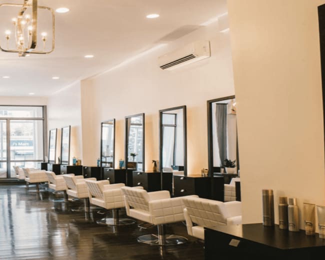 The image shows a modern hair salon with several styling stations, each equipped with mirrors and white chairs, lined along a well-lit interior.