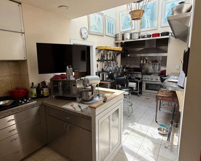 The image shows a kitchen with stainless steel appliances, a central island with a mixer, and various cooking utensils hanging on the walls.