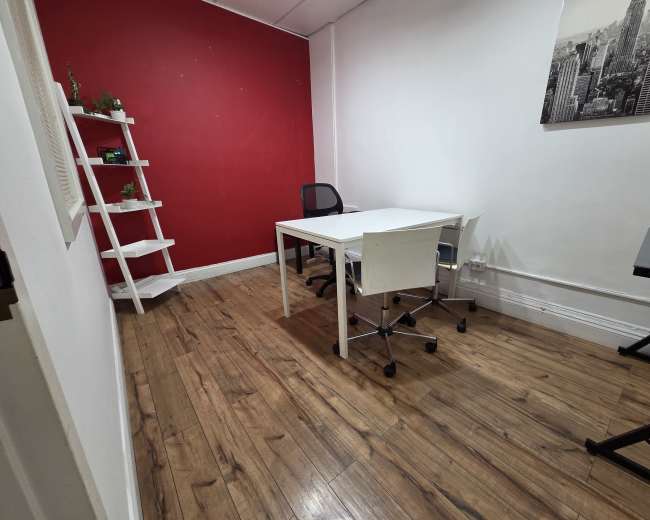 The image shows a small office space with a red accent wall, a white table, two chairs, and a shelf with plants.