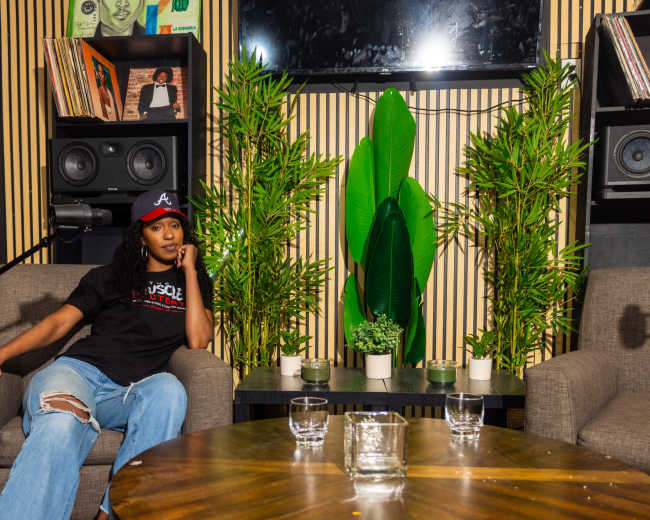 A person sits on a couch in a stylish room decorated with plants, speakers, and vinyl records.