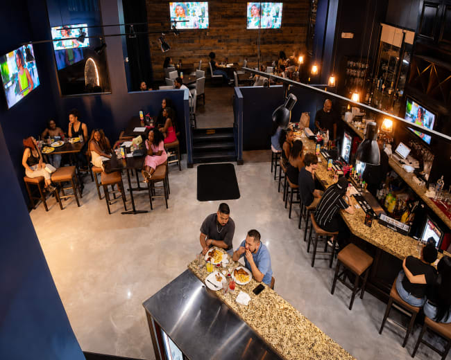 The image shows a vibrant restaurant interior with customers seated at tables and a bar, featuring televisions on the walls and a decorative ambiance.