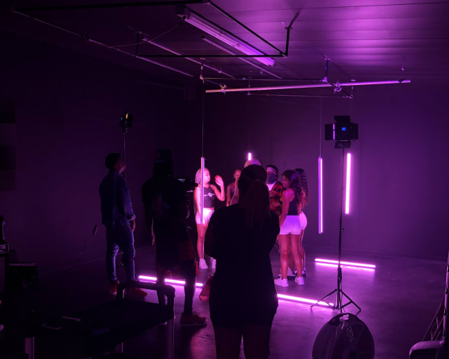 A group of people is gathered under neon lights in a studio setting, with a fan and equipment visible in the foreground.