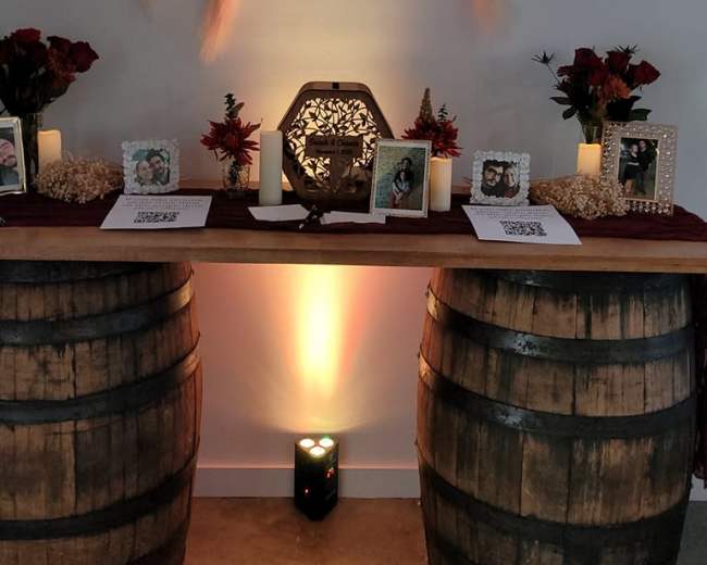 A decorated table with two wooden barrels as bases, featuring a floral arrangement above, framed photographs, candles, and a QR code display.