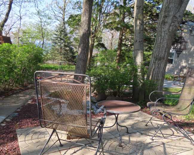A patio area features a metal chair and a small table surrounded by trees and shrubs.