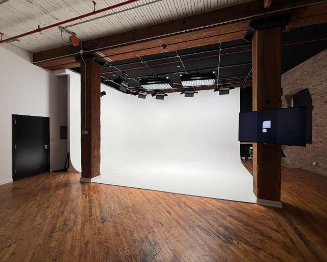 The image shows a spacious studio with wooden floors, exposed brick walls, and a bright, white backdrop, featuring overhead lights and a monitor on a side wall.