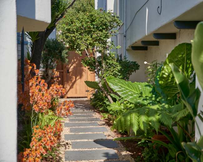 A stone pathway leads through lush greenery and vibrant flowers to a wooden door set against a white wall.