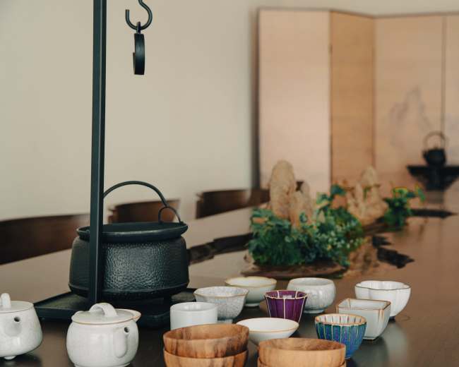The image shows a long wooden table set with various tea vessels and bowls, alongside a decorative arrangement of small rocks and greenery.