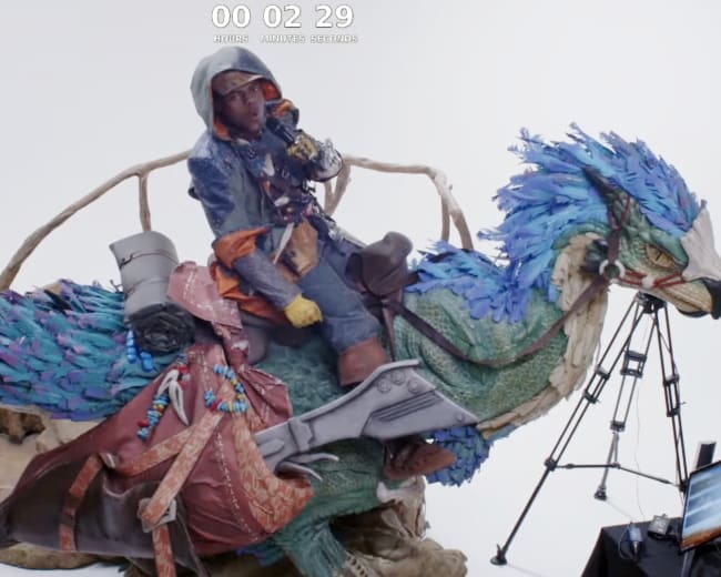 A person dressed in colorful attire sits atop a large, intricately detailed bird statue in a studio setting, with cameras set up nearby.