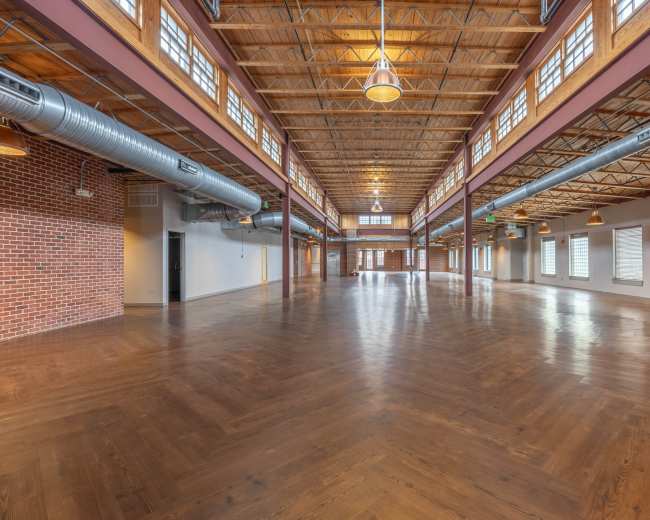 The image shows a spacious, well-lit interior of a large room with wooden flooring, high ceilings, and large windows.