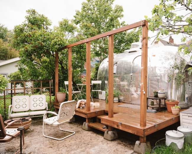 A silver Airstream trailer next to a wooden deck surrounded by greenery and patio furniture.