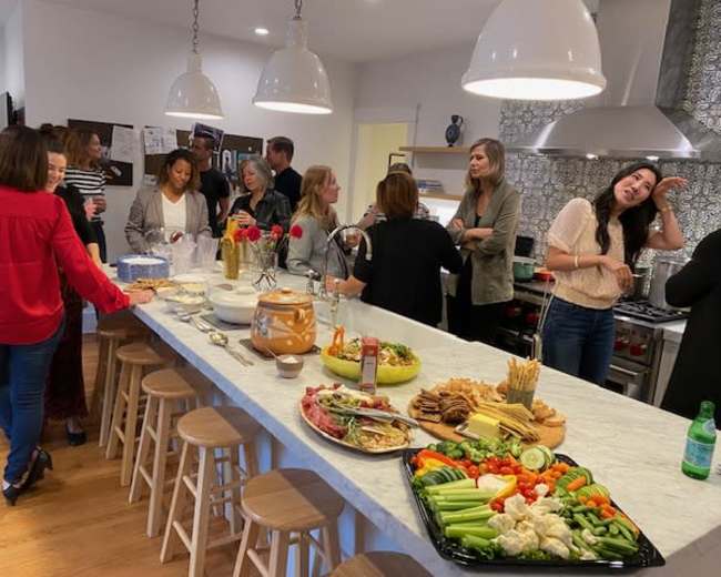A group of people gathers in a modern kitchen, socializing around a large marble island adorned with various trays of vegetables, meats, and snacks.